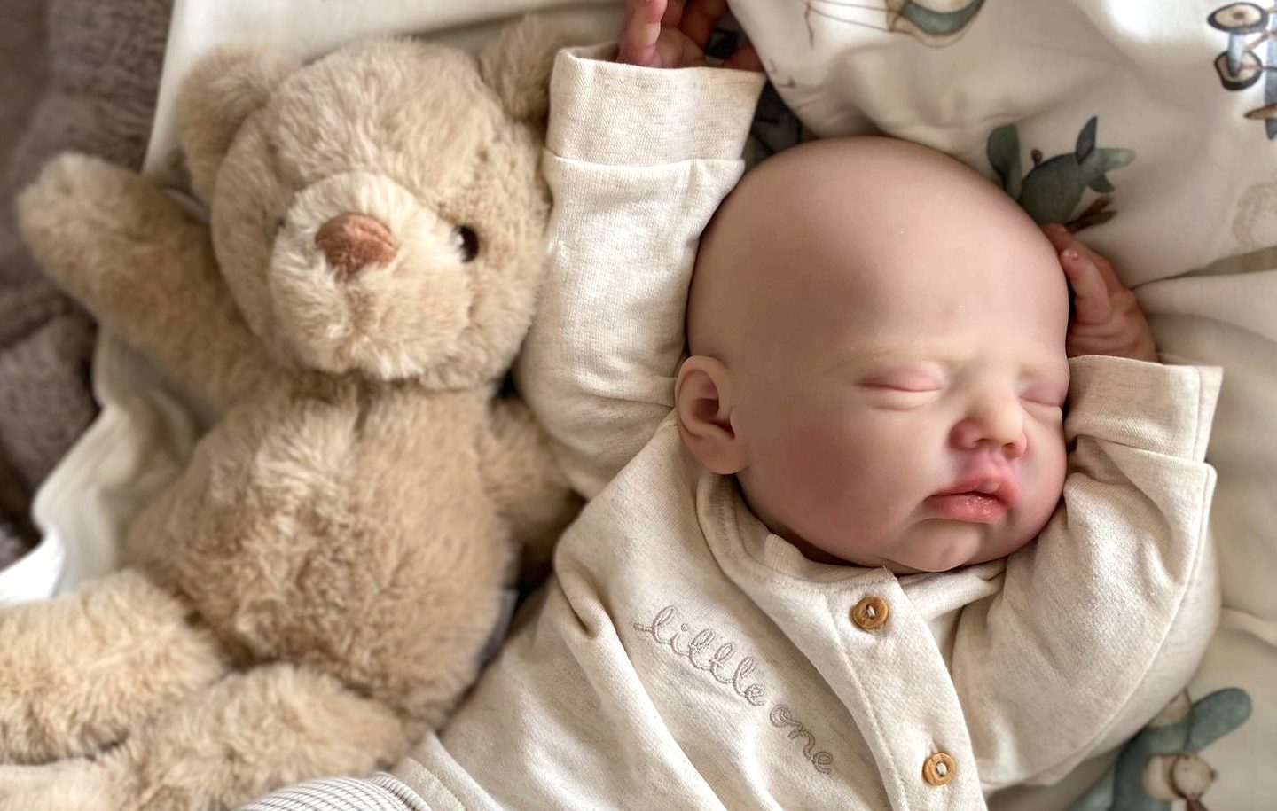 A reborn baby doll asleep in a cream romper, next to a teddy bear on a patterned blanket.